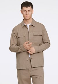 Light brown button-up jacket with two chest pockets, long sleeves, and a straight hem. Worn over a white t-shirt. Smooth texture.