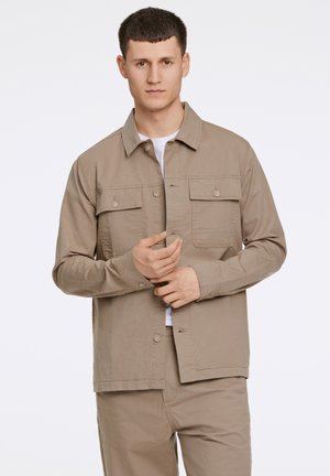 Light brown button-up jacket with two chest pockets, long sleeves, and a straight hem. Worn over a white t-shirt. Smooth texture.