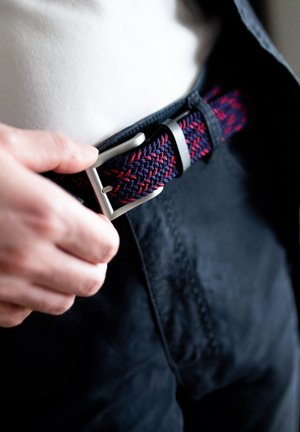 Woven elastischer Gürtel in Marineblau mit roten Akzenten, ausgestattet mit einer silbernen Metallschnalle. Hand, die den Gürtel an dunklen Hosen justiert.