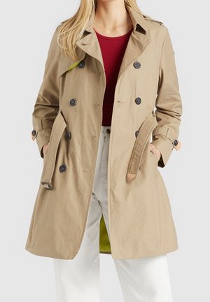 Beige dobbeltradet trenchcoat med sorte knapper og bælte, båret over en rød top og hvide bukser, hænder i frakkens lommer, forfra.