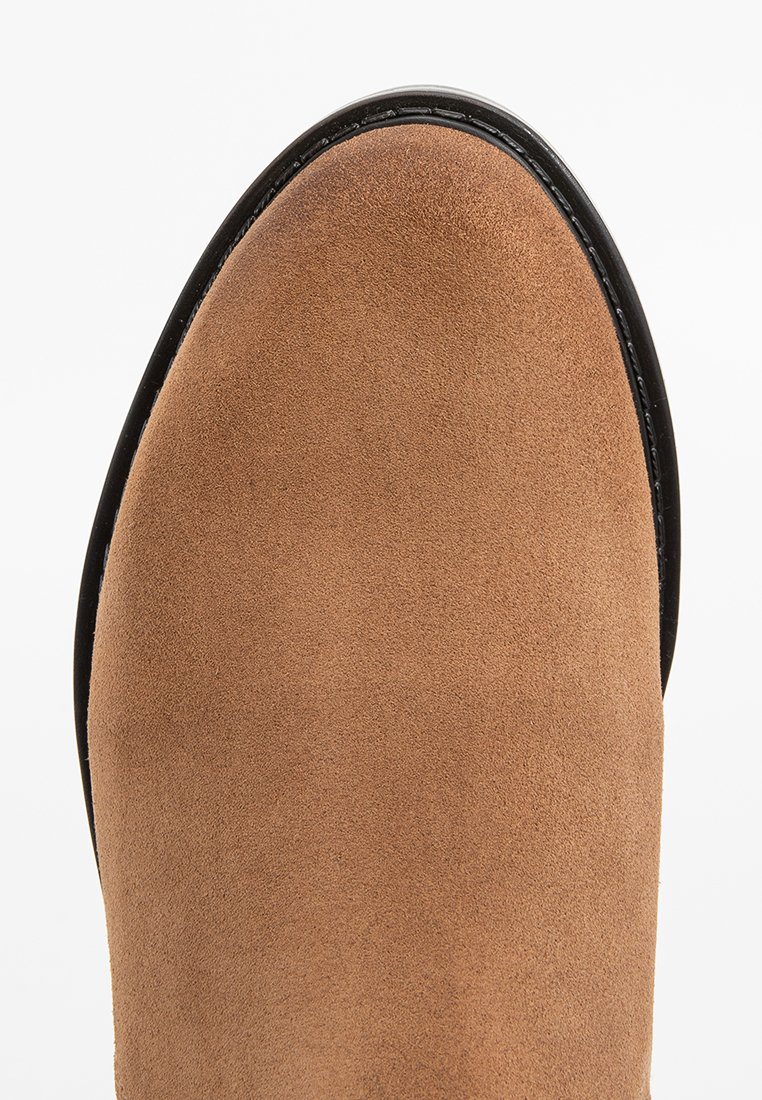 Bota de ante en tono marrón claro con punta redondeada, que presenta un borde negro suave a lo largo de la suela. Textura suave y diseño limpio.