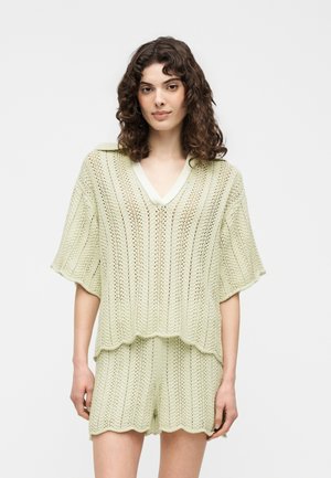 Kvinde med krøllet hår iført lys grøn strikket V-hals top med vide ærmer og matchende strikkede shorts, stående mod hvid baggrund.