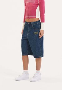 Blue denim knee-length shorts with a yellow embroidered logo on the front pocket, paired with white sneakers and a pink knit top.
