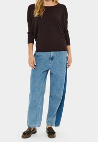 Femme portant un haut à manches longues marron foncé, un jean en denim bicolore bleu, et des mocassins marron, debout devant un fond blanc uni.