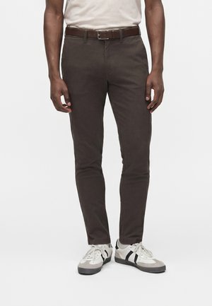 Homme portant un pantalon moulant marron foncé, une ceinture en cuir marron, des baskets blanches avec des rayures noires et une chemise blanche rentrée.