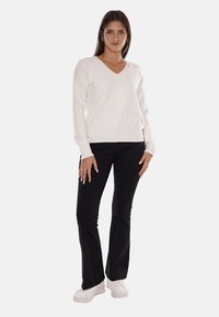 Maglione bianco a scollo a V realizzato in maglia morbida, abbinato a pantaloni neri attillati. La modella indossa sneaker bianche ed è in piedi su uno sfondo neutro.