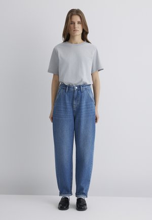 Helle graue Baumwoll-T-Shirt, lockere Passform; hochgeschnittene, weite blaue Jeans; knöchellang mit leichtem Verblassen; kombiniert mit schwarzen Schuhen.