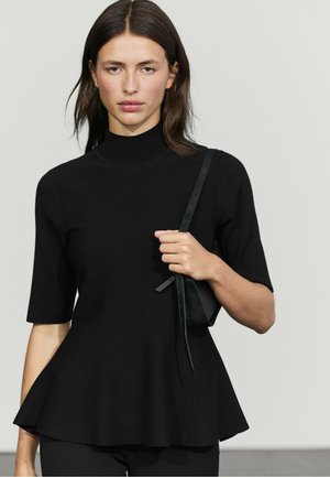 Donna con capelli scuri fino alle spalle che indossa un top peplum nero a maniche corte, tenendo una tracolla nera per borsa sulla spalla.