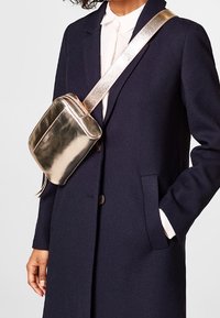 Borsa a tracolla metallica argento con chiusura a zip, forma rettangolare e tracolla regolabile, indossata sopra un cappotto blu scuro.