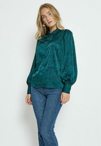 Satinbluse in smaragdgrün mit floralem Muster, Knopfleiste und puffärmeln, kombiniert mit blauen, eng anliegenden Jeans.