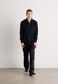 Camisa de pana negra con botones y un bolsillo en el pecho, combinada con pantalones negros y zapatos negros, sobre un fondo liso.