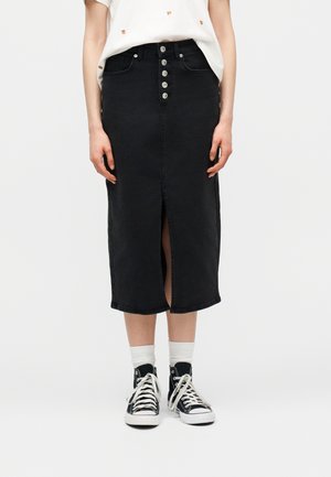 Žena má na sebe čiernu rifľovú sukňu do kolien s prednými gombíkmi a rozparkom, biele ponožky, čierne vysoké tenisky a biele tričko.