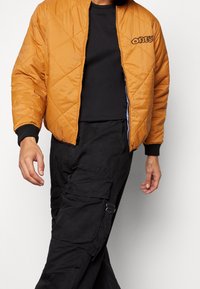 Veste bomber matelassée jaune moutarde avec des poignets côtelés noirs et un logo brodé, portée sur un t-shirt noir col rond et un pantalon cargo.