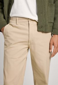 Pantaloni in cotone beige con chiusura a pulsante anteriore, davanti piatto e tasche laterali. Abbinati a un cardigan verde strutturato.