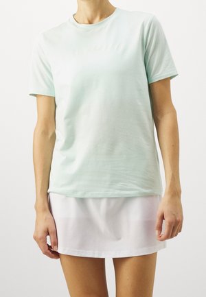 Henkilö, jolla on päällään vaalean mintunvihreä lyhythihainen T-paita ja valkoinen minihame, seisoo yksiväristä valkoista taustaa vasten.