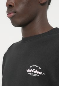 Κοντινό ενός ατόμου που φοράει μαύρο φούτερ με λευκό κείμενο λογότυπο «Definition of Jack & Jones Est. 1990 Strong and Authentic Quality Denim» στο στήθος.