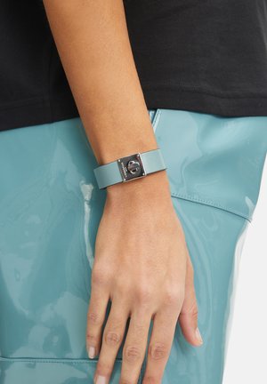 Bracelet en cuir bleu clair avec un fermoir rectangulaire en argent et des détails en métal, porté sur un poignet au-dessus d'un vêtement teal brillant.