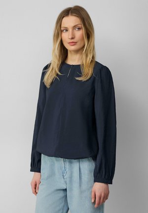 Dunkelbluse mit langen Ärmeln aus strukturiertem Stoff, rundem Ausschnitt und einem zentralen Reißverschluss. Besitzt geraffte Ärmel und eine lockere Passform.