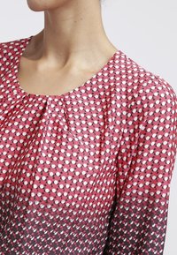 Blusa rossa e rosa con scollatura rotonda. Presenta un motivo a pois di dimensioni variabili, tessuto fluido e dettagli plissettati sulla scollatura.