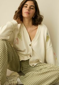 Creme cardigan med blomsterbroderi og knapper, parret med grønne gingham bredbenede bukser og hvide sneakers. Glat stoftekstur.