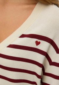 Long-sleeve, V-neck sweater in cream with maroon stripes; features small red heart embroidery on the left shoulder; soft cotton texture.