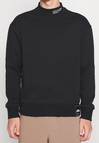 Sudadera negra de cuello redondo con mangas largas, puños y dobladillo de canalé, etiqueta de tela en el cuello, sutil marca en la manga. Textura suave.