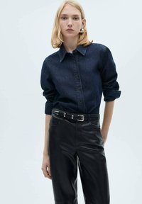 Camicia in denim blu scuro con chiusura a bottoni e maniche arrotolate, abbinata a pantaloni in pelle neri a vita alta con cintura.