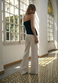 Svart strapless topp, vita vida byxor, stående mot stora fönster med dekorativt kakelgolv. Mjukt naturligt ljus belyser scenen.