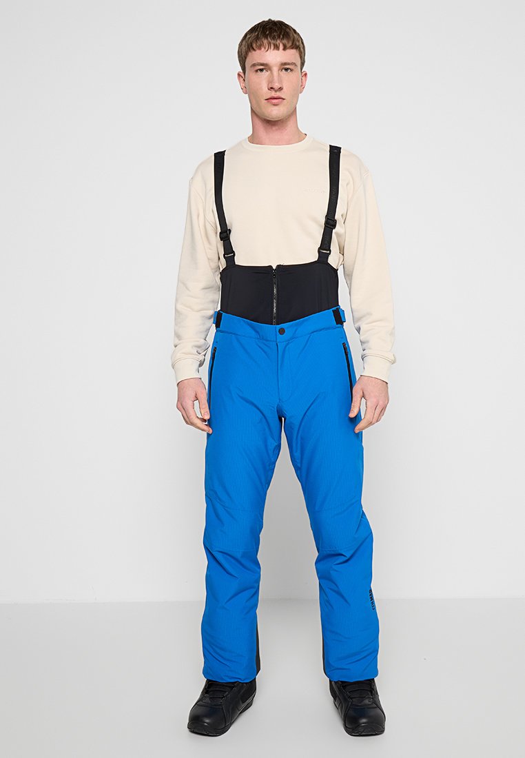 Niebieskie spodnie narciarskie z regulowanymi czarnymi szelkami i zasuwanymi bocznymi kieszeniami. Materiał ma teksturowane wykończenie i posiada zamek błyskawiczny z przodu.