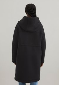Czarny długi płaszcz z kapturem wykonany z miękkiego materiału. Posiada luźny krój, boczne kieszenie oraz prosty, bezszwowy design bez widocznych zapięć.