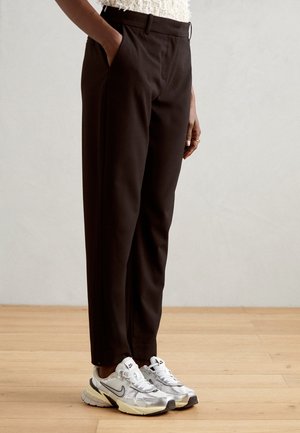 Osoba nosząca dopasowane ciemnobrązowe spodnie z bocznymi kieszeniami i białe sneakersy Nike, stojąca na jasnej drewnianej podłodze na tle neutralnej ściany.
