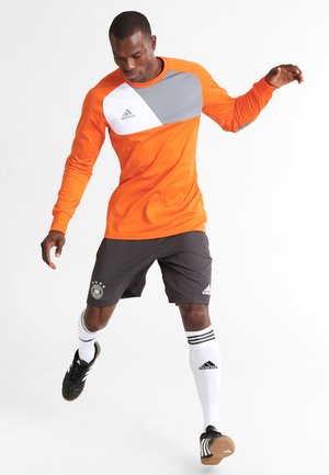 Orange long-sleeve athletic shirt with gray and white accents, paired with black shorts featuring a logo, white socks, and black cleats.