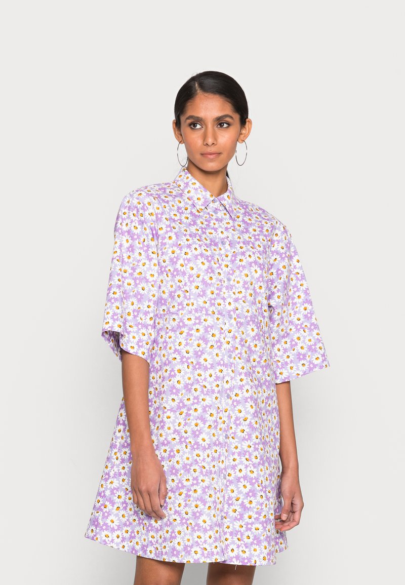 Vestido camisa de manga curta em tecido lilás claro com estampa de margaridas, apresentando um colarinho com botões e um corte descontraído.