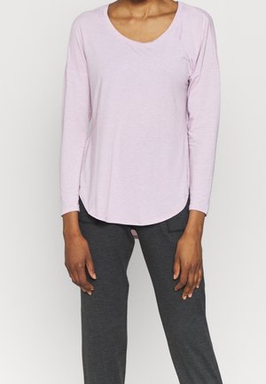 Woman wearing a light purple long-sleeve shirt with a rounded hem and dark gray pants standing against a plain background.