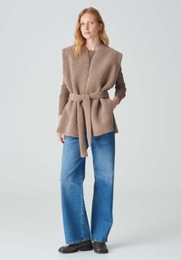 Beige, strukturierten Weste mit Wickeldesign und Gürtel, kombiniert mit blauen geraden Jeans und schwarzen Stiefeletten. Minimalistischer Hintergrund.