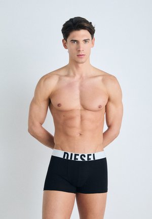 Boxeri negri cu un elastic lat alb pe care este inscriptionat logo-ul "DIESEL". Țesătura pare moale și se mulează pe corp. Modelul stă cu brațele în spate.