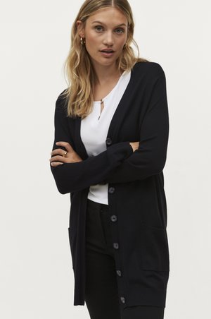 Cardigan blu marina realizzato in materiale lavorato a maglia, con chiusura a bottoni, due tasche anteriori e polsini a costine. Indossato sopra una maglietta bianca.
