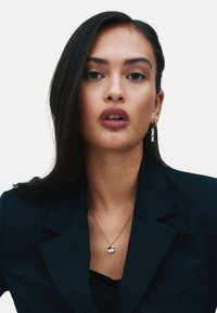 Gold pendant necklace with a clover design, worn with a dark blazer. The earring features the word "DREAM" also in gold.