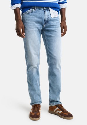 Light blue denim jeans with a straight cut. Worn with brown sneakers featuring textured suede and white stripes, paired with a blue striped sweater.