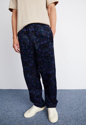 La personne porte un pantalon ample bleu foncé à motif floral, une chemise beige à manches courtes et des baskets blanches, debout sur un tapis gris.