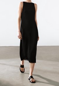 Robe midi sans manches noire, en tissu lisse, coupe décontractée, avec une fente sur le côté, associée à des sandales noires plates avec une large lanière et un détail texturé.