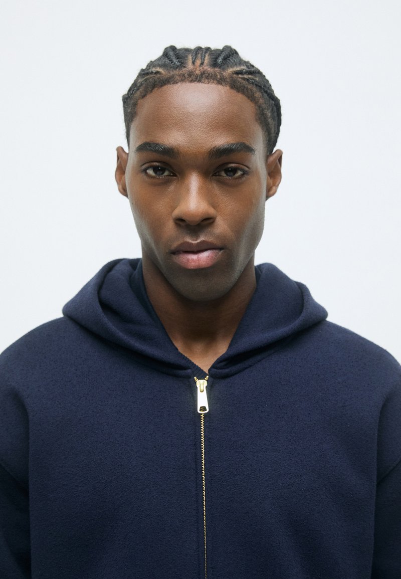 Marineblauwe hoodie met een rits aan de voorkant, voorzien van een gouden rits. De stof lijkt zacht met een gestructureerde afwerking en heeft een ontspannen pasvorm.