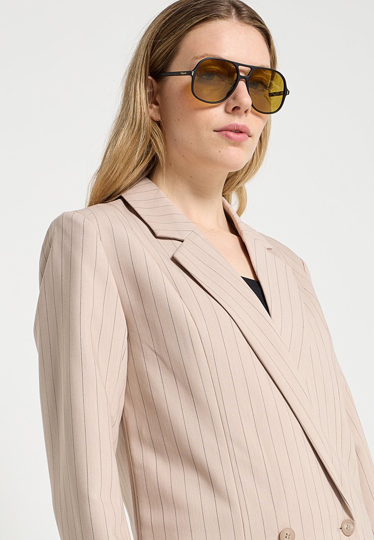Femme portant un blazer beige à rayures et de grandes lunettes de soleil teintées, faisant légèrement face à droite, sur un fond clair uni.