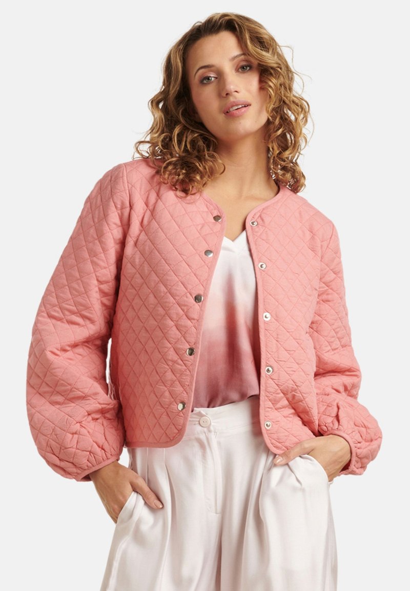 Roze gewatteerde jas met een ronde halslijn, zilveren drukknoopjes en pofmouwen. Gedragen over een lichte blouse en witte broeken.