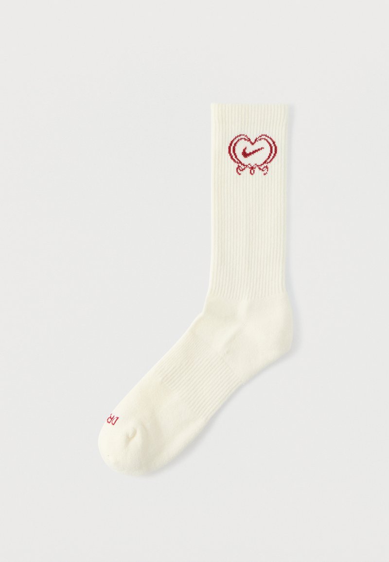 Calzino a coste color crema con grafica di un cuore rosso, decorato con una striscia curva e un fiocco in cima, con marchio rosso vicino alla punta.