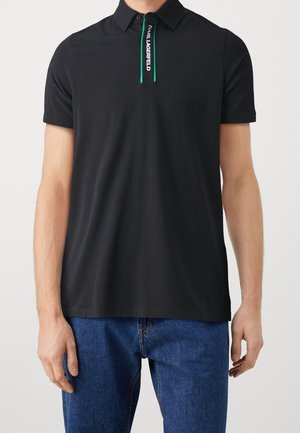 Uomo che indossa una polo nera a maniche corte con cerniera bordata di verde e scritta bianca "KARL LAGERFELD", abbinata a jeans blu.