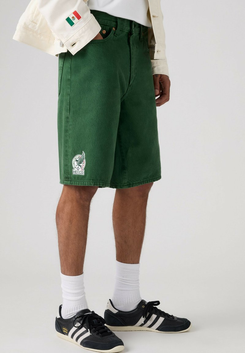 Person trägt grüne Shorts mit einem Mexiko-Emblem, weiße Socken, schwarze Adidas-Sneaker und eine leichte Jacke mit einem mexikanischen Flaggen-Patch am Ärmel.