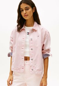Giacca di jeans rosa con bottoni argentati, maniche arrotolate che rivelano un rivestimento a righe. Abbinata a un crop top bianco e pantaloni rosa chiaro.