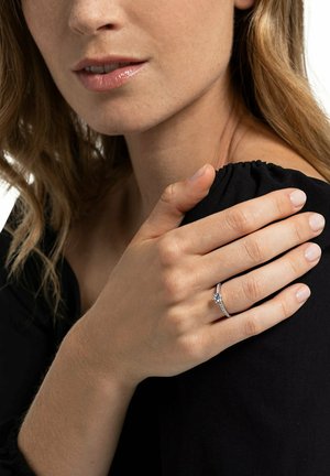 Frau mit hellem Teint und welligem Haar, die sanft ihre Schulter berührt und einen silbernen Verlobungsring mit einem einzelnen Diamanten trägt.