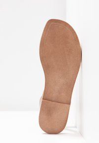Semelle d'une chaussure en daim marron avec une adhérence texturée au talon et à l'avant, positionnée à la verticale contre un mur d'angle blanc et le sol.